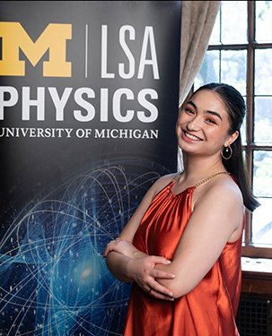 Nicole is smiling with her arms crossed in front of a large University of Michigan Physics poster.