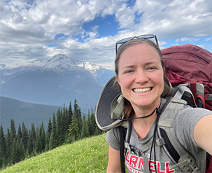 A selfie-style picture of a smiling woman with sunglasses on her head and an expedition backpack strapped on her back and chest. A snow topped mountainscape and lush, green rolling hills fill the background. The sky is a brilliant blue sky, smeared with white puffy clouds.