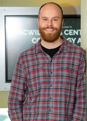 Brendan O'Connor is posed for this photo, leaning against a wall on his left side while wearing a red and gray plaid, flannel shirt.